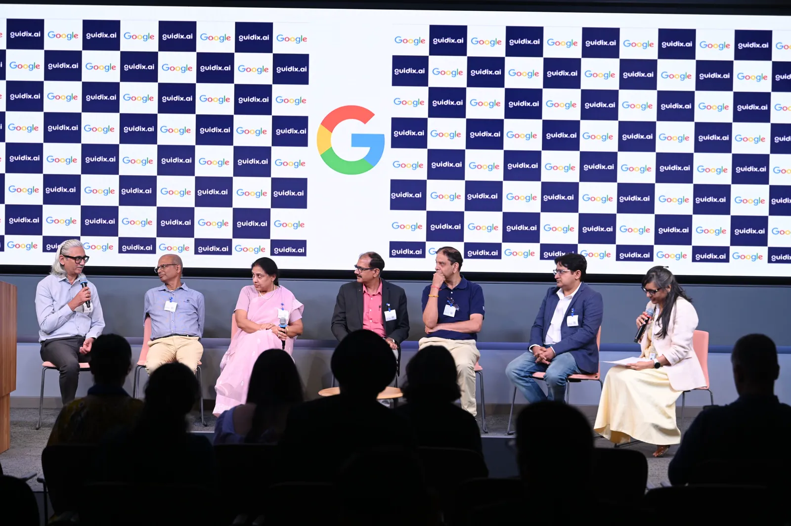 A panel of seven speakers on stage, seen from the audience, under the Google and guidix.ai backdrop
